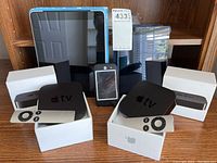 Two Apple TV units with remotes and boxes, two iPads with protective cases, and one iPhone smartphone, all placed on a wooden shelf.