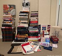 Wide view showing a large assortment of stacked books, paperbacks, hardcover books, pamphlets, and brochures on a carpeted floor. Books have various subjects including politics, history, and self-help.