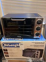 DeLonghi convection toaster oven on top of original box. Oven has stainless steel finish and three knobs on the right side.