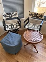 Two director chairs with artistic fabric seats and backs, a blue fabric pouf, and a wooden game table with carved chessboard surface on a hardwood floor.
