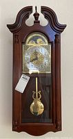 Frontal view of the Howard Miller wall clock showing wooden case, finial, clock face with numbers, moon phase dial and pendulum inside glass door.