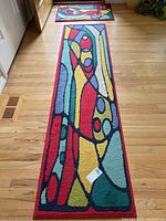 Photo of a long rectangular runner rug with colorful abstract design featuring red, yellow, green, blue, and purple sections with black outlines on a light wooden floor.