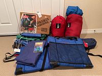 Full overview of lot showing The North Face sleeping bags in red sacks, Coleman camp stove box, various blue camping bags and accessories on beige carpet.