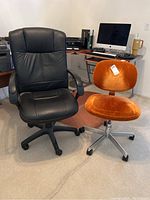 Two office chairs side by side in a home office with a computer desk in the background, one black leather and one orange velvet.
