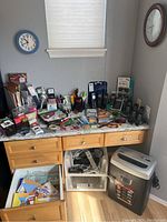 Wide view of desk with assorted office supplies including calculators, scissors, clip holders, paper shredder, and desk drawer open showing papers and envelopes.