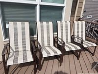 Four outdoor patio chairs with brown metal frames and striped beige-brown fabric upholstery on the deck against house windows.
