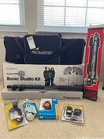 Full lot showing Promaster SystemPro Basic Studio Kit box with two light stands and softboxes, Manfrotto tripod box, black softbox carry bag, extension cord, flash trigger and photo paper spread on floor.