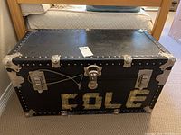 Front view of black travel trunk labeled 'COLE' with metal reinforcements and locking mechanism.