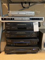 Stack of 6 VHS and DVD players from various brands including Sharp, Panasonic, Magnavox, and Symphonic on metal shelving.