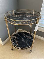 Front angled view of vintage two-tier bar cart showing gold metallic frame with decorative swirls and black marble-patterned shelves