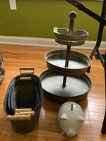 Three-tiered galvanized metal tray, ceramic piggy bank, and three metal planters with wooden handles arranged on hardwood floor.