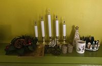 Five brass electric candle holders with white plastic tapers, plugged in with cords coiled in front, accompanied by a faux floral wreath on a gold plate, a decorative holiday tree, and a ceramic cup with Santa and reindeer design.