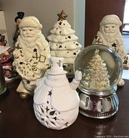 Overview of the lot showing two white ceramic Santa figures with gold highlights, a white ceramic Christmas tree, the musical snow globe with silver base, and the lighted snowman in front.