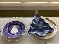 Six Christmas themed plates displayed including round plate with snowman, Christmas tree-shaped plate, and holly/pinecone motif plates