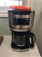 Front view of Hamilton Beach drip coffee maker showing stainless steel body, glass carafe with black handle, and coffee maker top with coffee filters packed on top.