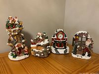 Four Christmas-themed figurines on a table, including a clock and snow-covered holiday scenes