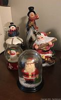 Photo shows two snow globes featuring Santa and snowman figures, a 9-inch snowman figurine, and a two-piece Santa figurine playing a musical instrument with chip on lid.