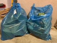 Two blue plastic bags filled with green floral oasis blocks on a concrete floor next to a wall.