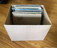 Cardboard box containing approximately thirty vinyl LP records upright on hardwood floor
