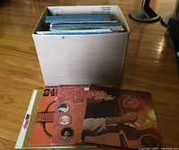 Photo of cardboard box containing approximately 40 LP record albums, showing some of the album covers at the top, including John Denver, country music titles, and a multicolored 24-track Sunshine label album.