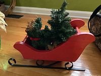 Side view of red wooden sleigh with decorative Christmas tree and two twig reindeer inside, showing black metal runners on bottom.