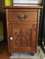 Front view of the small wooden side table showing drawer with ornate metal handle and carved wood cabinet door with white knob. Crack noted in carving.