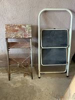 Photo showing two step stools side-by-side against wall on concrete floor. The left stool has paint-splattered wooden top and grooved wood steps with metal frame. The right stool is a white tubular metal folding stool with black rubber tread steps branded Tricam.