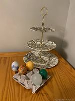 Three-tier dessert tray showing gold-tone risers and scenic black and white Royal Mail pattern with colored accents, standing on wooden surface, alongside five decorative eggs including one on a small metal stand.