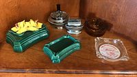 Multiple ceramic ashtrays, a trinket box with yellow flower decoration, and a clear glass ashtray with branding on a wooden shelf.