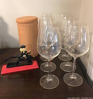Eight tall clear wine glasses with thin stems and wide bases arranged in two rows on a dark wooden surface next to a tall terracotta clay wine chiller with a lid and a vintage black metal nutcracker holding a red cloth.