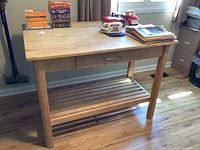 Front and diagonal views of the wood butcher block work table with a flat top, single drawer, and slatted lower shelf. Shows foldable drop-down leaf in closed position.