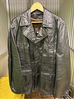 Front view of black leather vintage jacket on hanger showing collar and pocket details.