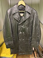 Front view of a black leather men's trenchcoat jacket on a hanger, showing double-breasted button front and wide lapels.