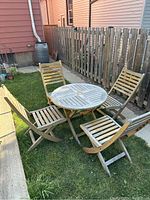 Full view of vintage teak outdoor dining set with round table and four folding chairs arranged on grass near a wooden fence; shows weathering and aged grey coloring of wood