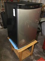 Front angled view of Danby bar fridge on wooden stand showing stainless steel door and black sides.