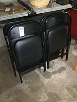 Four black folding chairs stacked folded with black vinyl padded seats and backrests, black metal frames.