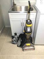 Front view of Bissell Revolution carpet cleaner standing upright with cord wrapped, two bottles of cleaning solution and black accessory bag on floor
