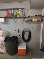 Green trash can with black car mats rolled on top, next to an orange and white cooler, in garage setting.