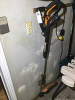 Full view of Worx cordless string trimmer leaning against wall showing orange and black color scheme and shaft.