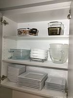 Photo of cupboard shelves with stacked white square plates and bowls, glass bakeware dishes including CorningWare casserole with floral pattern and glass loaf pans, measuring cup and bowl.