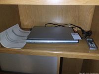 Magnasonic DVD player on wooden shelf with remote and manual beside it