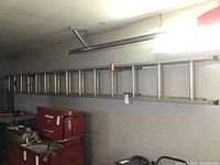 Full length side view of the aluminum extension ladder hung on a wall above red toolboxes and other items.