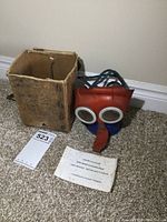 Overall view of vintage child's gas mask standing next to old cardboard box on carpeted floor with instruction leaflet partially visible