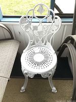 Front view of white cast metal outdoor chair showing the ornate floral and vine backrest and circular lattice seat pattern.