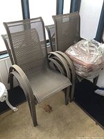 Four metal frame outdoor patio chairs with gray mesh fabric stacked, with four round beige cushions wrapped in plastic on top, minor wear visible on carpet and chairs.