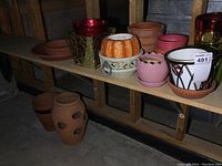 Photo showing a shelf holding a variety of mostly clay and ceramic planters of different colors and styles including red, orange, pink, white with patterns, and natural clay color planters. Includes round and openwork designs.