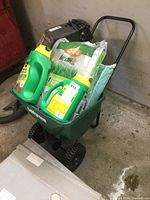 Green Golfgreen lawn spreader filled with two jugs of Shake N Feed fertilizer and several bags of lawn care products and soil, placed on concrete floor.