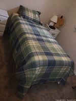 Single bed made up with plaid quilt and pillow, bedside table with lamp visible