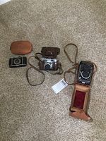 Three vintage cameras including Voigtlander, Foth Derby, and a small leather pouch on carpeted floor