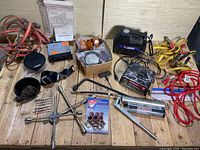 Various automotive tools and accessories laid out on a wooden surface including jumper cables, battery charger, lug wrench, grease guns, car horn, mirrors, lug nuts and assorted items.